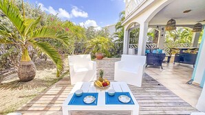 A covered outdoor seating area with white chairs and a table set for two, surrounded by tropical plants and a wooden deck.