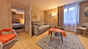Un salon chaleureux avec un canapé, une petite table, un tapis à motifs et une vue sur une chambre à coucher, visible à travers une porte ouverte.
