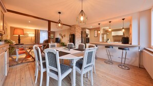 Un espace repas avec une table et des chaises blanches, une cheminée en pierre et des lampes suspendues.