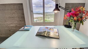 A room with a large window, a table with a book and a lamp, and a vase with flowers. - Paradise View, Maison créole avec piscine privée à 5 min d`Orient Bay (Orient Bay)
