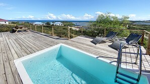 A wooden deck with a swimming pool, lounge chairs, and a view of the ocean and distant buildings. - Paradise View, Maison créole avec piscine privée à 5 min d`Orient Bay (Orient Bay)