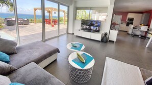 A spacious living area with a large TV, a sofa, and a dining area with a kitchen in the background.