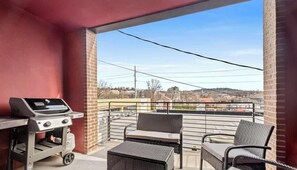 A balcony with a grill, wicker chairs, and a view of buildings in the distance.