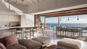 Eine moderne Küche mit weißen Schränken und einer Bar, eine gemütliche Sitzecke mit Sofa und Hocker sowie ein Essbereich am Pool mit Blick aufs Meer.