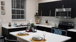 A modern kitchen with a central island, black cabinets, stainless steel appliances, and a window with blinds. - Tropical Pardise A (Tampa)