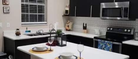 A modern kitchen with a central island, black cabinets, stainless steel appliances, and a window with blinds.