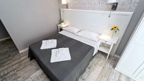 A neatly made bed with white linens and a dark gray bedspread, two bedside tables with lamps, a patterned wallpaper, and a wooden floor. - Standard double room (Roma)