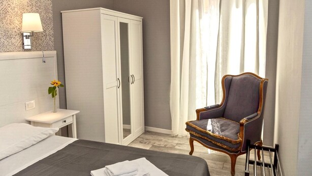A hotel room with a bed, a wardrobe, a chair, a bedside table, and a lamp. - Standard double room (Roma)
