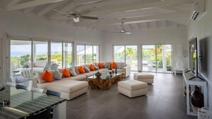 Ein geräumiges Wohnzimmer mit einem großen Glastisch, weißen Sofas mit orangefarbenen Kissen und einem Deckenventilator.