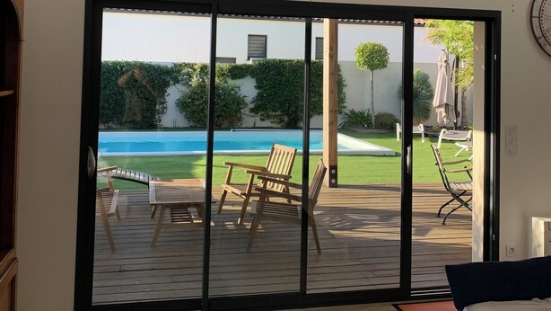 Una habitación moderna con un amplio ventanal corredera que da a una terraza exterior con piscina, sillas de descanso y vistas a un edificio de fachada blanca y vegetación.