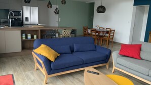 Una sala moderna con un sofá azul, un sofá gris, una mesa de centro de madera y un otoman amarillo. Hay una zona de cocina con microondas, refrigerador y armarios.