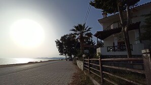 A paved pathway beside a road with a wooden fence and a building with a balcony. - Zennova 22 Nikiti Sea Side House (Sithonia)