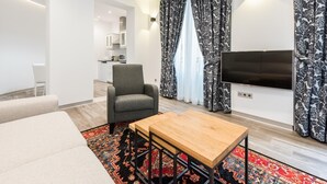 Ein modernes Wohnzimmer mit einem grauen Sessel, einem Couchtisch aus Holz und einem an der Wand montierten Fernseher. Im Hintergrund ist eine Küche mit weißen Schränken und einer Mikrowelle zu sehen. Das Zimmer verfügt über bunte Vorhänge mit Muster und einen farbenfrohen Teppich.