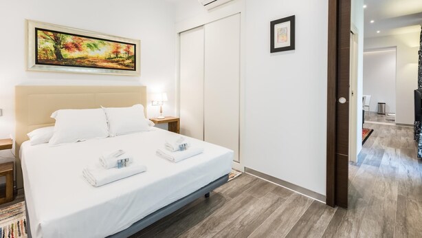 Ein modernes Hotelzimmer mit einem Bett, einem Nachttisch, einem Gemälde, einer Klimaanlage und einer Tür zu einem weiteren Raum.