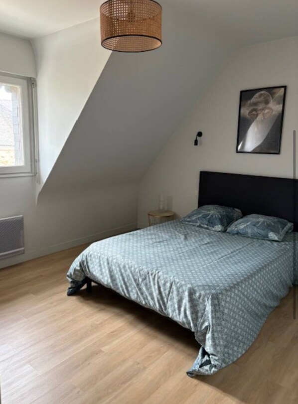 Un dormitorio con techo inclinado, una cama con ropa de cama estampada, una lámpara fijada a la pared y un cuadro enmarcado.
