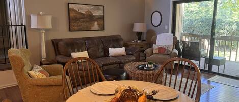 A living room with a brown leather sofa, a round dining table with placemats, and a large window with a view of greenery.