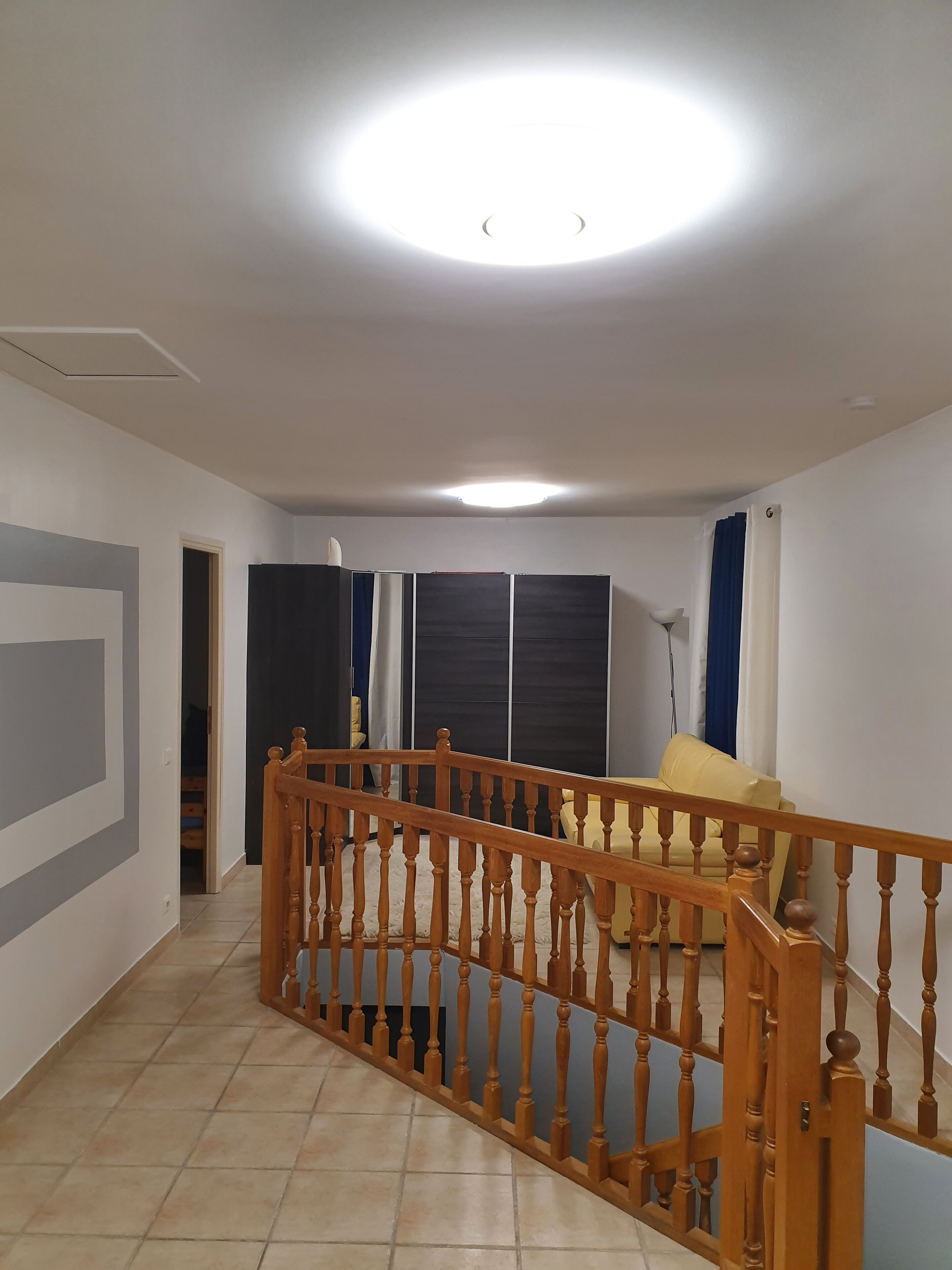 Een kamer met een houten trap, een plafondlamp, een bed met een wit hoofdeinde en een raam met blauwe gordijnen.