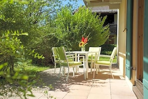 An outdoor patio area with a table and chairs, surrounded by greenery and a building entrance.