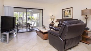A modern living room with a leather sofa, a flat-screen TV, a coffee table, and a dining area with chairs and a glass door leading to a balcony. - Villa Sanibel 2B (Sanibel)