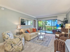 A living room with a beige sofa, patterned armchair, and wicker chairs. A small round table with a glass top is in the center. A large window offers a view of the outdoors. - Sand Pointe 212 (Sanibel)