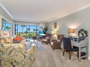 A spacious living room with a large window overlooking palm trees, a dining area, and a cozy seating arrangement. - Sand Pointe 212 (Sanibel)