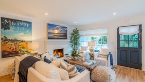 A living room with a fireplace, a large poster of a beach, and a door with a window. - Casa Miramar - Coastal Farmhouse Chic Getaway (Santa Barbara)