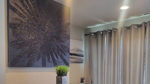 Een eethoek met een glazen tafel, twee krukken, een wanddecoratie en een raam met gordijnen.