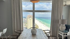 A dining area with a wooden table, chairs, and a hanging light fixture. A large window offers a view of a beach and ocean.