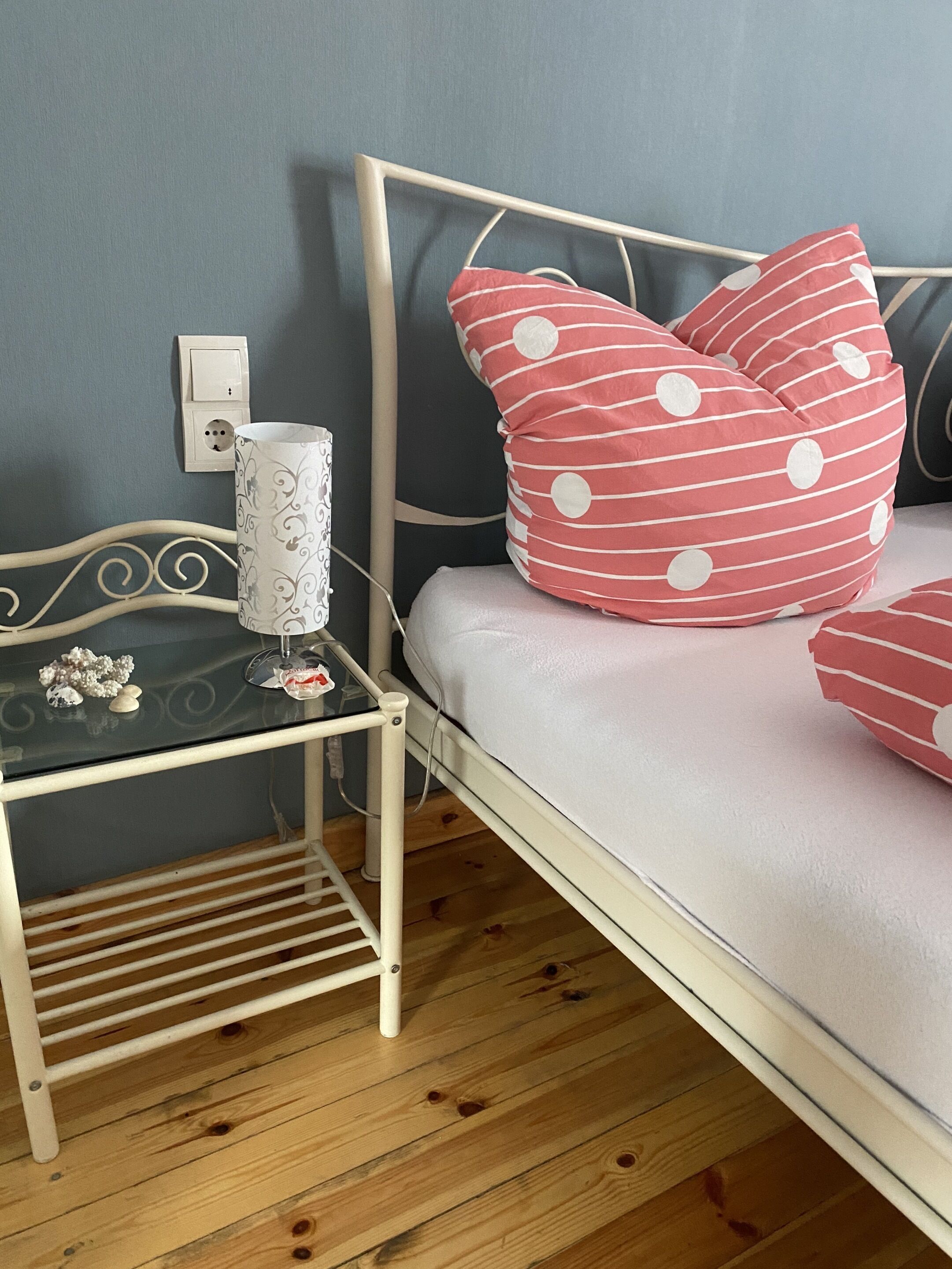 Un lit avec un cadre blanc et des oreillers à pois rouges, une table de chevet avec un plateau en verre et une prise électrique murale.
