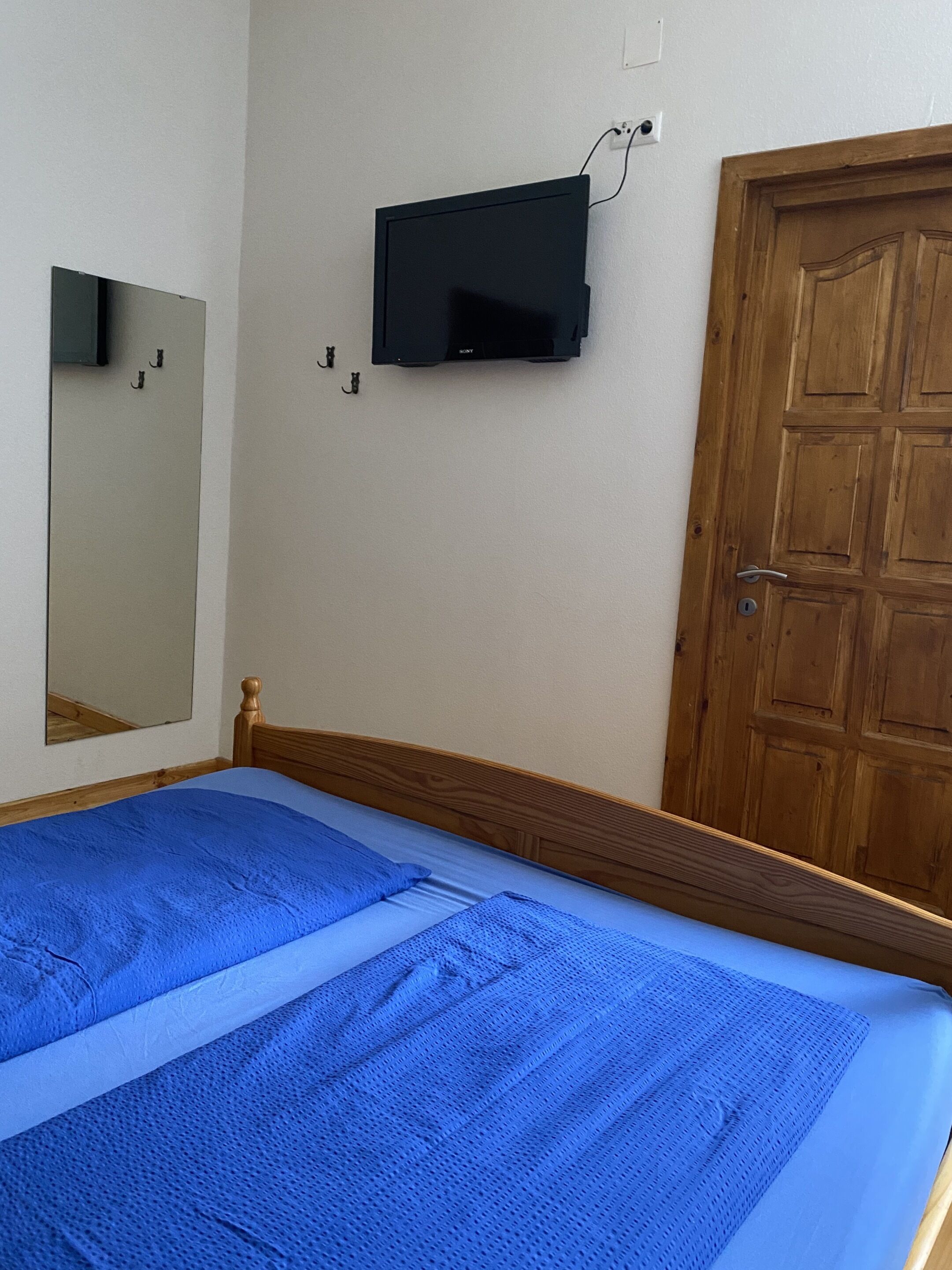 Un dormitorio con cama, una puerta de madera, televisión montada en la pared y un espejo.