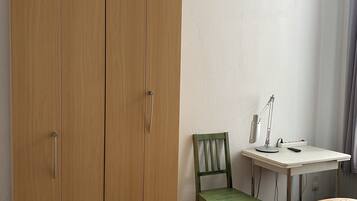 A bedroom with a wooden wardrobe, a green chair, a desk with a lamp, and a bed with colorful pillows.