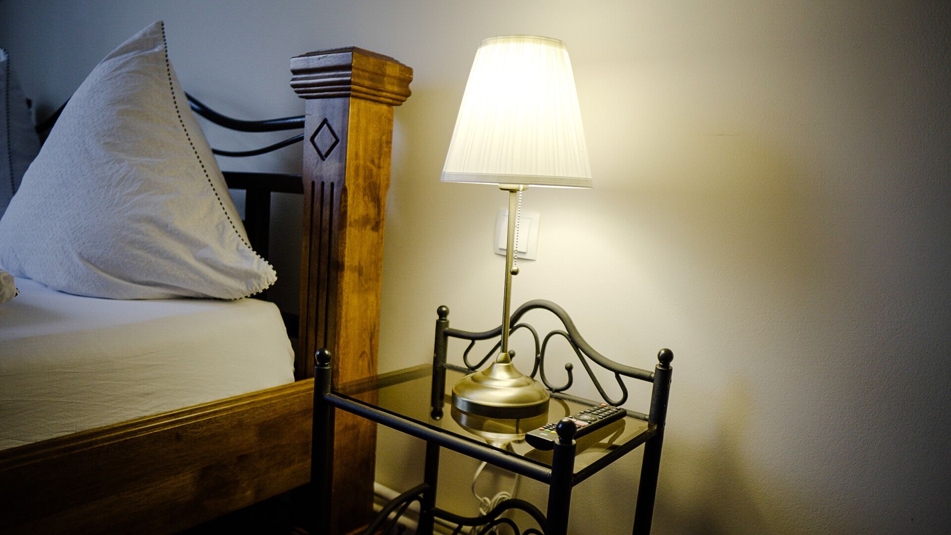A bed with a white pillow and a bedside table with a lamp and a remote control.