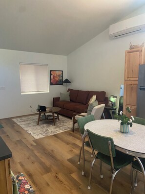 Ein Wohnzimmer mit einem braunen Sofa, einem runden Tisch mit Stühlen, einem Fenster mit Jalousien und einem Holzboden.