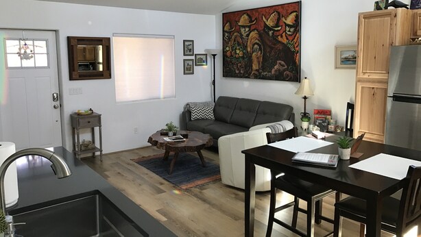 Una sala de estar moderna con un sofá gris, una mesa de centro y un comedor con mesa y sillas de madera. Hay una zona de cocina con un lavaplatos y un refrigerador.