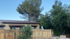 Una casa con cerca de madera, un jardín con plantas y rocas, y un árbol grande al fondo.