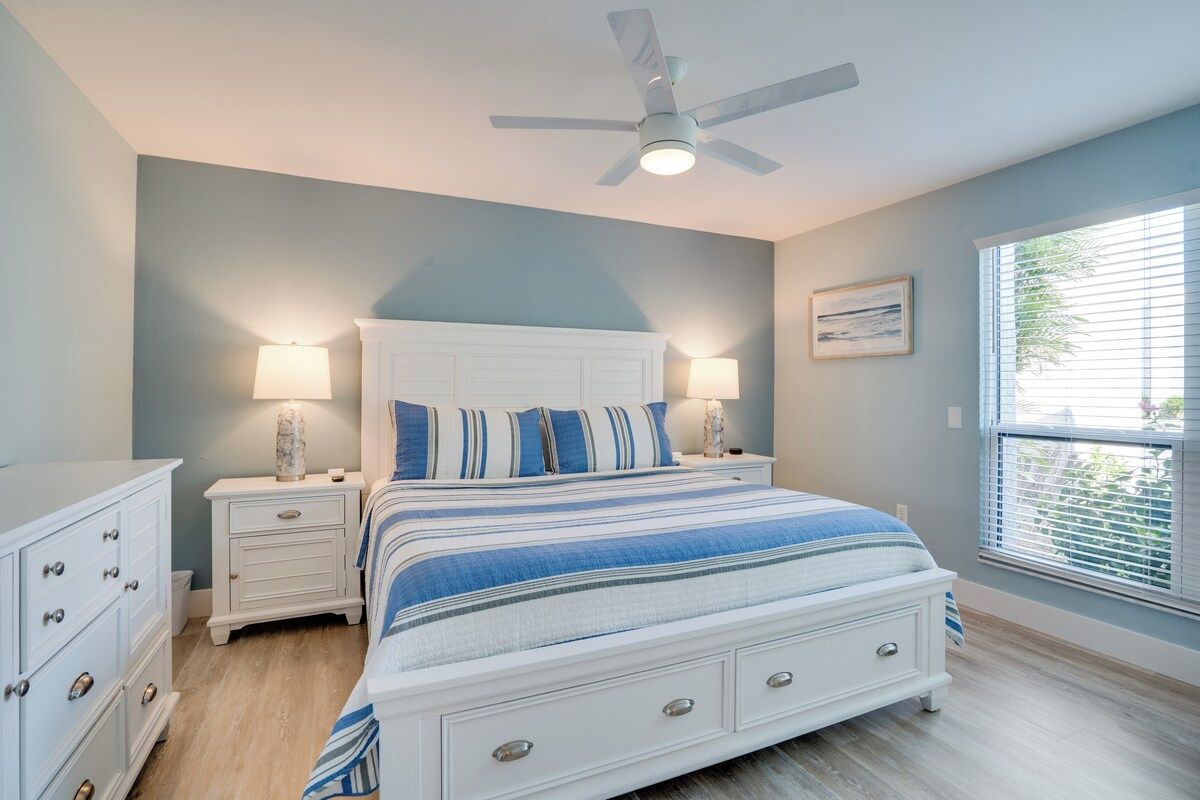 Une chambre à coucher avec un lit, des tables de chevet, un ventilateur de plafond et une fenêtre avec des stores.