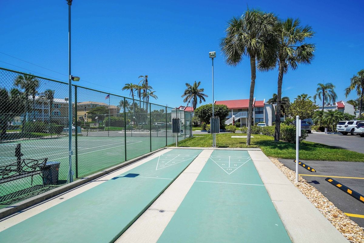 Una cancha de shuffleboard verde al aire libre con una cerca, palmeras y un edificio con techo rojo al fondo.