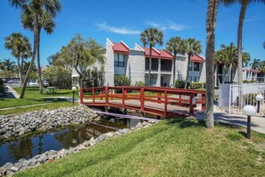 A red bridge over a small water feature, surrounded by palm trees and a building with a red roof.