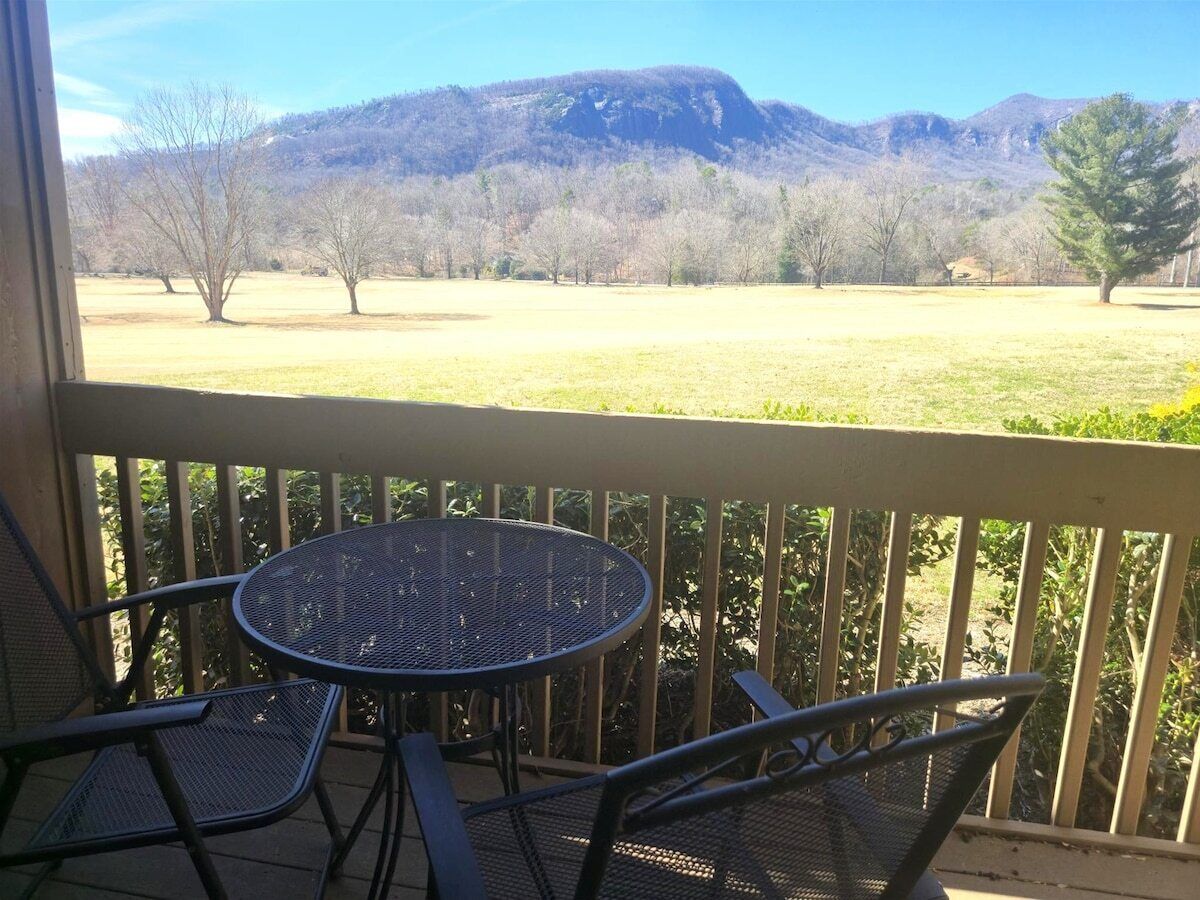 Varanda com mesa e cadeiras, com vista para um campo gramado e montanhas.