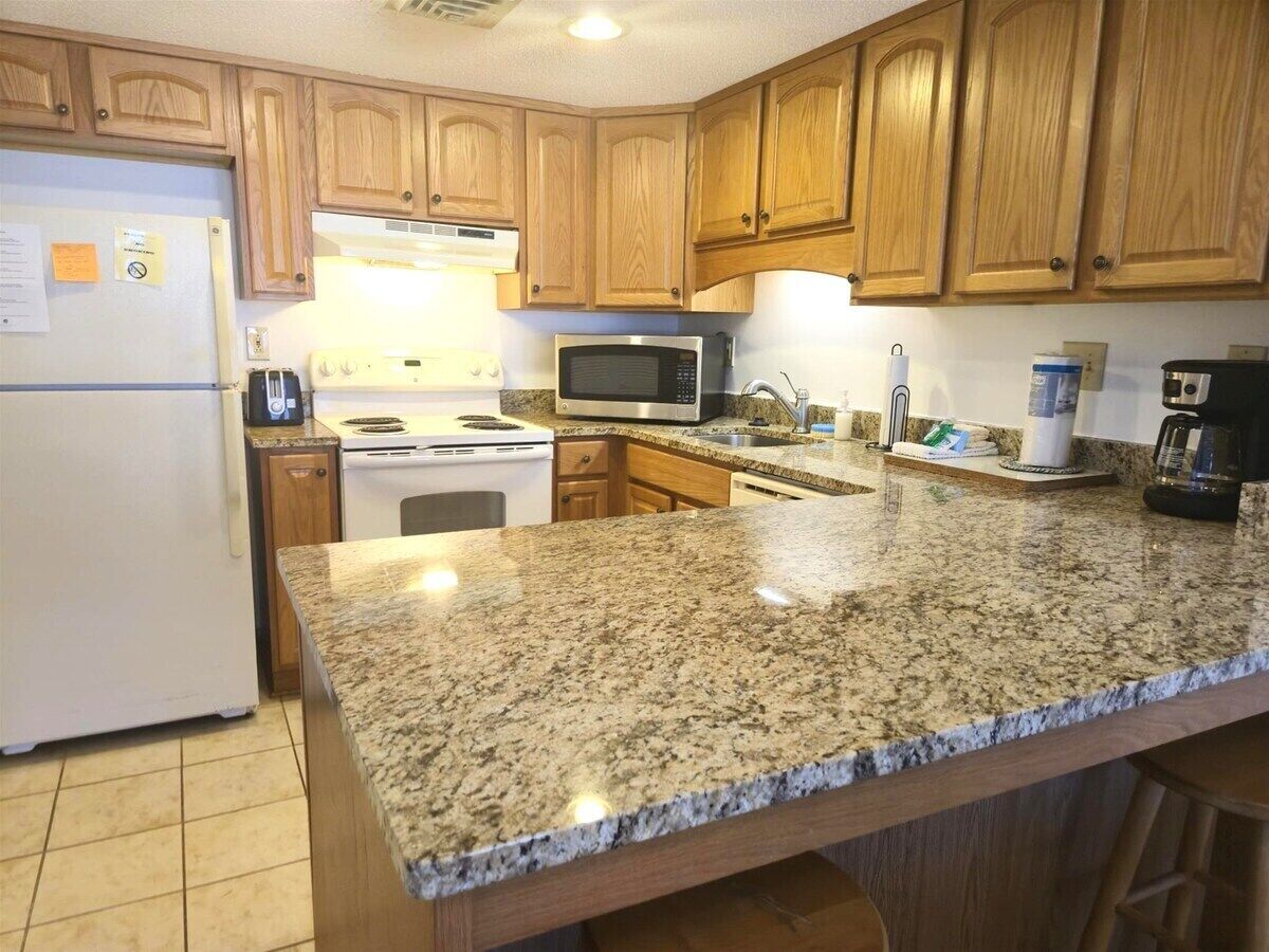 A kitchen with granite countertops, wooden cabinets, a refrigerator, a microwave, a stove, and a coffee maker.