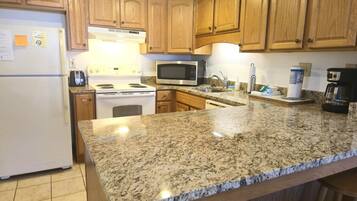 A kitchen with granite countertops, wooden cabinets, a refrigerator, a microwave, a stove, and a coffee maker.