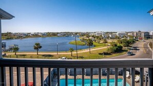 A view from a balcony overlooking a lake with a swimming pool, palm trees, and a road with cars. - Pool! First row from beach front with 2 Private Balconies (Carolina Beach)