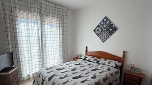 Een slaapkamer met een bed, een televisie op een standaard, een kamerkleed met dessin, een decoratief wandobject en een plafondventilator.