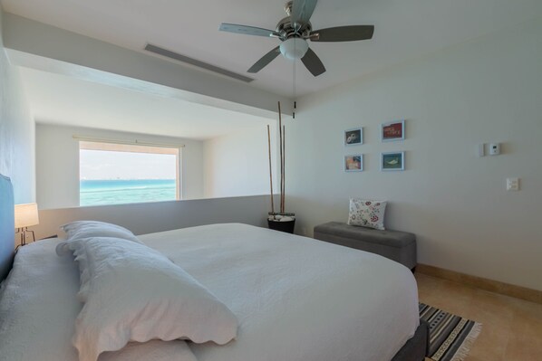 A bedroom with a large bed, a ceiling fan, a window with a view, a small table, and a gray sofa.