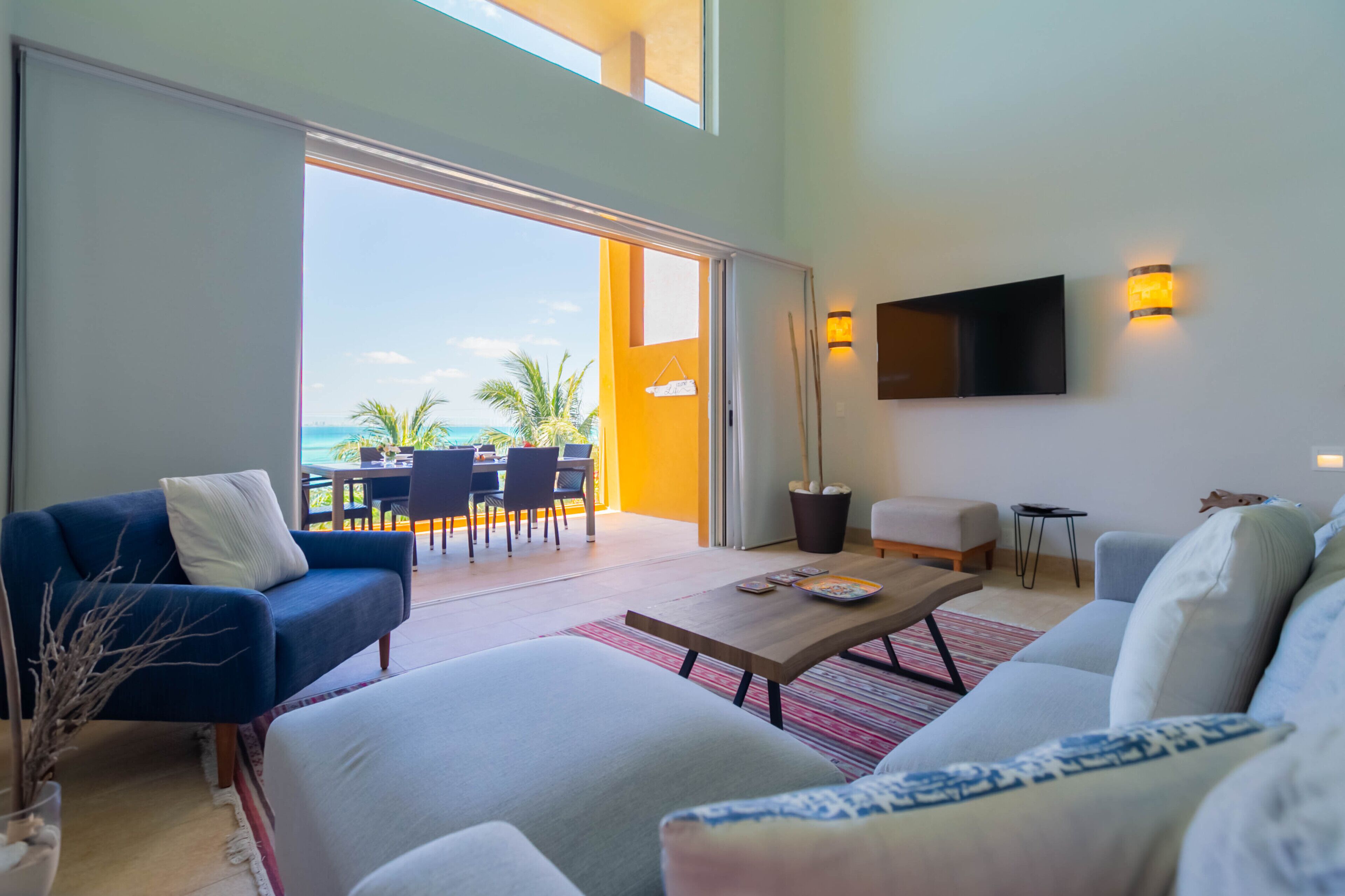 Una sala de estar moderna con un sofá grande, una mesa de centro y un televisor montado en la pared. Cuenta con un balcón con vistas al océano y palmeras.