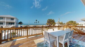 A wooden deck with a white table and chairs, overlooking a beach with palm trees and a clear sky.