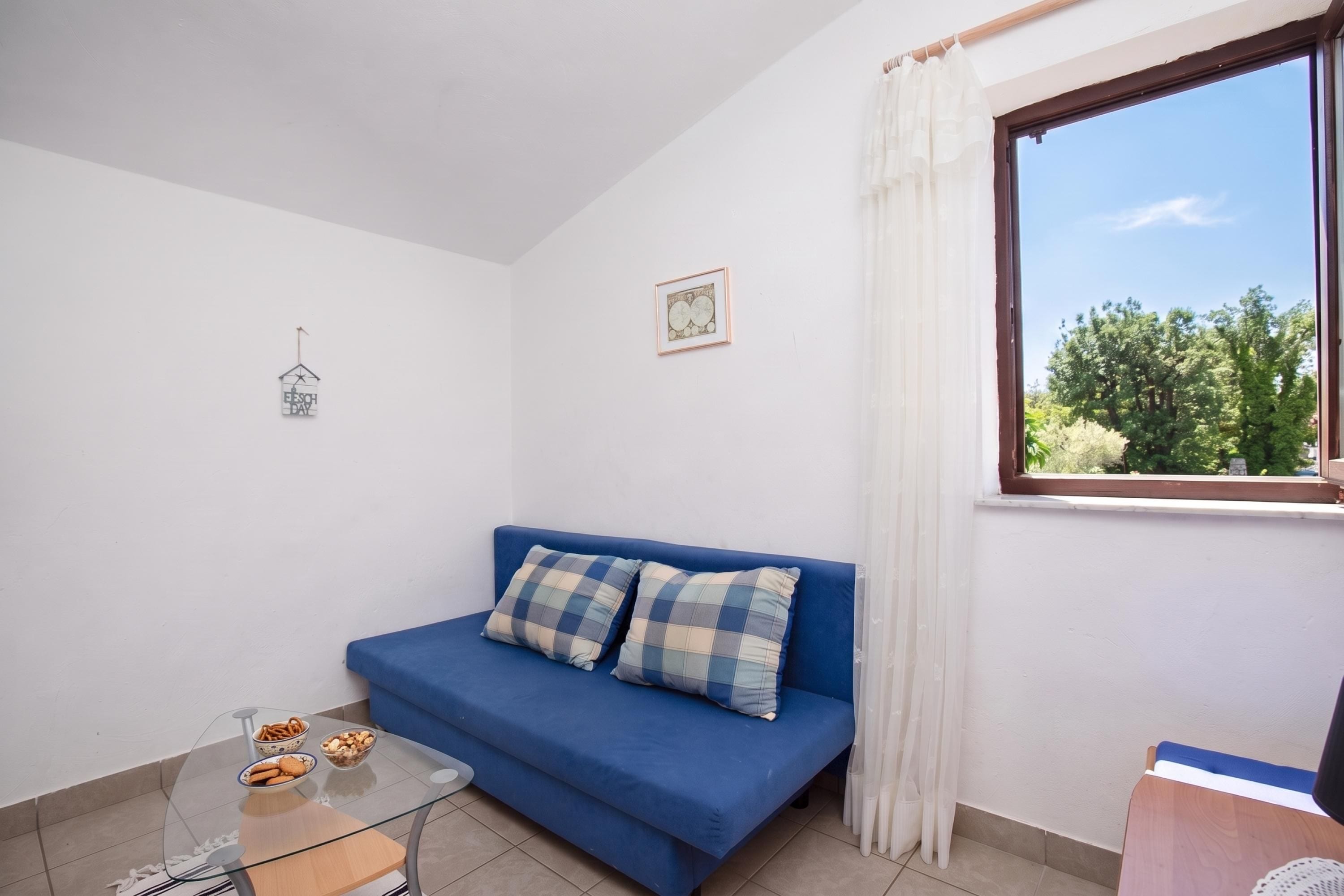 Una habitación con sofá azul, una mesa de vidrio con aperitivos, una ventana con cortinas y una pared con un cuadro enmarcado.
