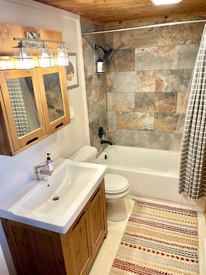 A bathroom with a white sink, toilet, and bathtub. It features stone tiles, wooden cabinets, and a shower with a handheld showerhead. - Dog Friendly•Close to Monarch•King Beds (Salida)