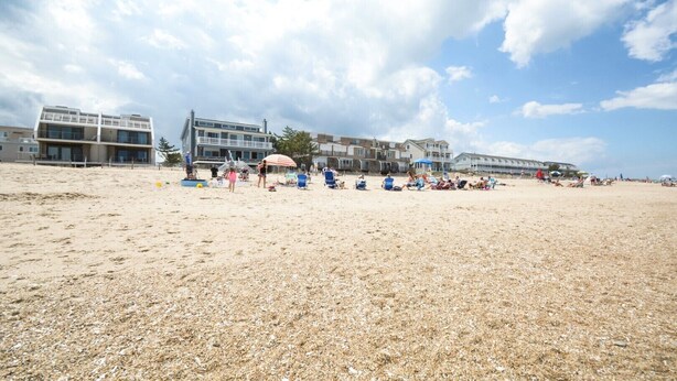 A beach with multiple buildings in the background, including a large structure with a balcony. - Beach home nestled between the beach and the bay (eastside) (Bethany Beach)