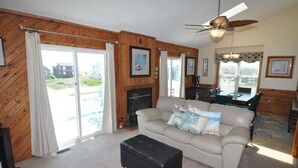 A living room with a sofa, ottoman, fireplace, ceiling fan, and large windows with curtains.